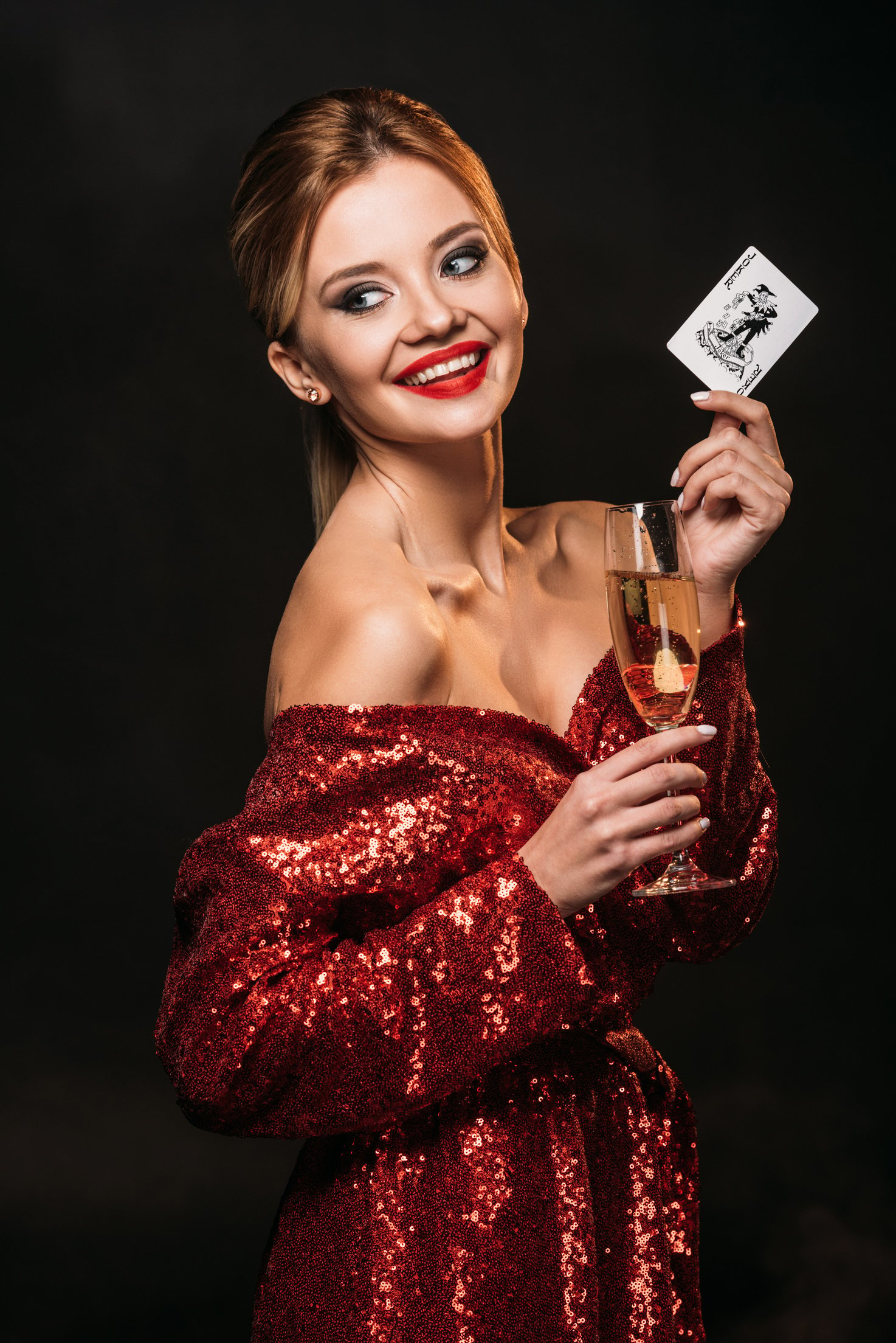 smiling attractive girl in red shiny dress holding joker card and glass of champagne isolated on black, looking away